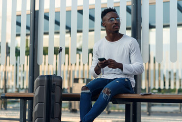 Young bearded man tourist with backpack and smartphone stand on public transport stop and waiting tram in modern city center. Travel concept.