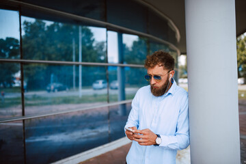 Fototapeta premium A young handsome caucasian businessman with sunglasses stands in front of the company and types a message on the phone