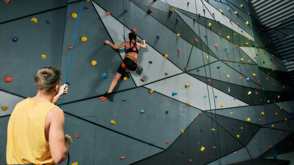 Male instructor holding rope, teaching, looking at woman in safety equipment and harness while she...