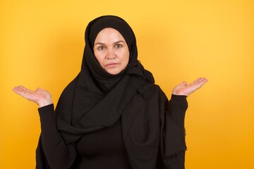 Hesitant Beautiful middle aged muslim woman wearing black hijab over yellow background shrugs shoulders, looks uncertain and confused. Have no answer