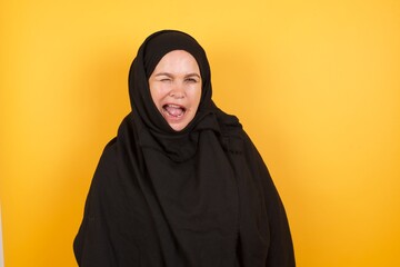 Beautiful middle aged muslim woman wearing black hijab over yellow background  winking looking at the camera with sexy expression, cheerful and happy face.
