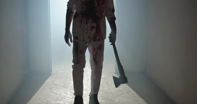 Bloodstained maniac walking in hallway of old house. Bloodthirsty butcher wearing white stained apron and carrying an axe, escaping after murder 4k footage