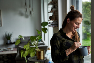 Smiling mid adult woman holding coffee mug while standing by window at home