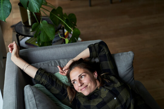 Smiling woman stretching hands looking away while lying on sofa at home