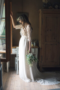 Young woman in elegant wedding dress holding bouquet looking out of the window