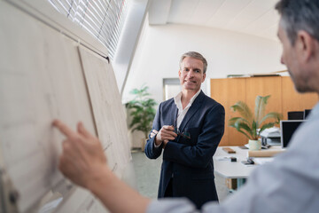 Confident mature businessman looking while listening to male colleague explaining strategy over whiteboard at office during meeting