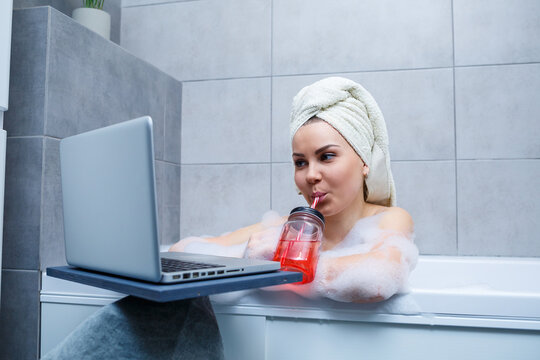 A young woman with a towel on her head drinks juice watching a movie on a laptop while sitting in a bath in a beauty salon. Relax in the bathroom without linen. Body care and relaxation concept.