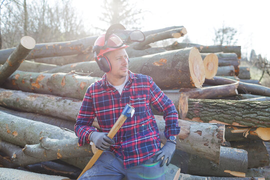 Thoughtful lumberjack holding axe while sitting on logs during sunny day