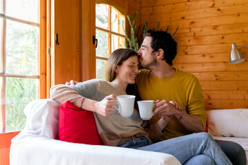 Romantic boyfriend holding coffee mug kissing on woman's forehead while sitting in log cabin