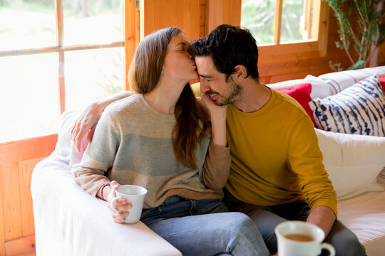 Romantic woman kissing on boyfriend's forehead while sitting over sofa in log cabin