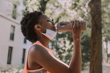 Sportive young woman with protective mask drinking water from flask