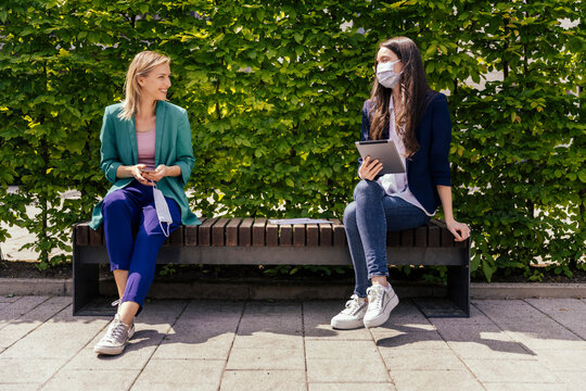 Two businesswomen sitting on bench outside and keeping their distance while wearing face mask