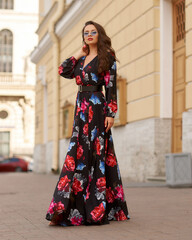 Fototapeta na wymiar Young beautiful lady wearing black dress with colorful floral design and standing at city street. Pretty woman outdoor full length portrait. Fashion model.