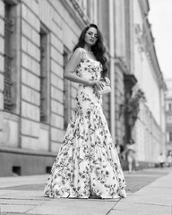 Young beautiful woman with long brunette wavy hair walking at city street. Elegant female wearing long colorful white and red floral dress. Girl in ball gown. Full length fashion portrait