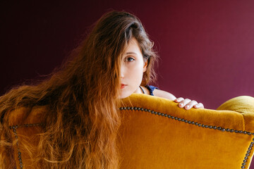 Close-up portrait of young woman with long brown hair leaning on golden chair against colored background