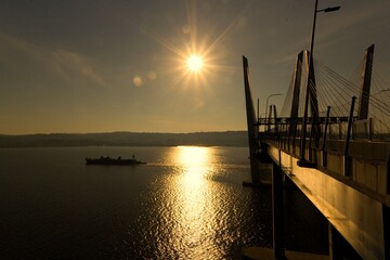 Obraz premium Tarrytown, NY / USA: A landscape image of sunrise at the Governor Mario M. Cuomo Bridge. A silhouette of a ship passing by the bridge, with Tarrytown in the background.