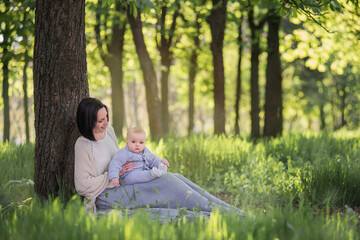 Fototapeta premium Modern beautiful young brunette mother with a short haircut, holding a toddler in a gray knitted romper in her arms. A girl and her son are sitting under a tree, playing together, hugging. Mom kisses 