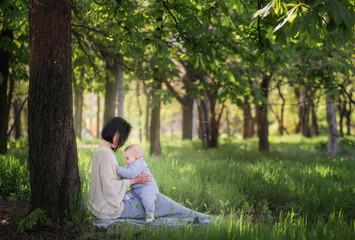 Modern beautiful young brunette mother with a short haircut, holding a toddler in a gray knitted romper in her arms. A girl and her son are sitting under a tree, playing together, hugging. Mom kisses 