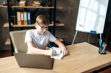 Distant learning. Stay home. Smart charismatic schoolboy with glasses is studying online, he is sitting at home at a table with a laptop and concentrated write information in a notebook