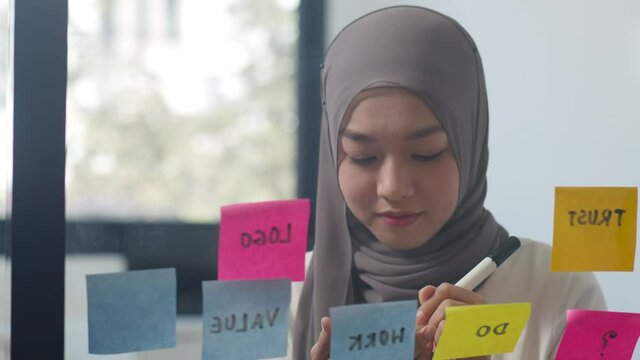 Asia muslim lady write information stick a sticky note on glass board in new normal office. Working from home, remotely work, self isolation, social distancing, quarantine for coronavirus prevention.