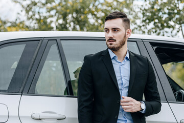Handsome young adult posing beside his white car in a black suit suit 