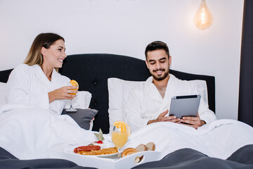 woman holding a glass of orange juice in bed while her partner is showing her something on his tablet, both laying in bed having breakfast
