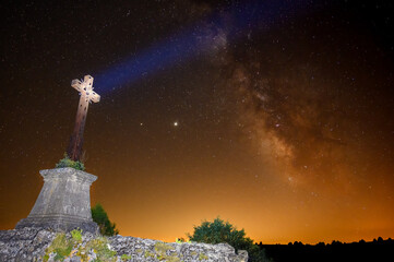 Apocalyptic image of a cross with an orange sky, the Milky Way behind and a beam of light impacting...