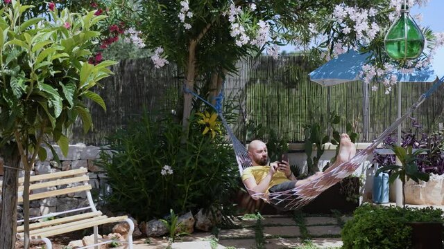 Bald guy writes messages on his smartphone and relaxes on the hammock in the garden outdoor