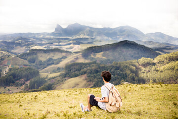 Fototapeta na wymiar Young adventurous traveler sitting on top of a mountain carrying a backpack and admiring the mountains of the State of Espirito Santo, Brazil. 