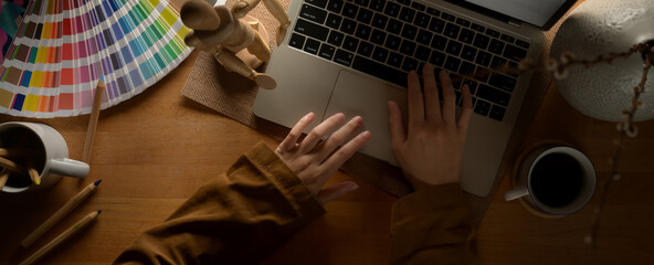 Female designer typing on laptop keyboard on worktable with supplies, painting tools and decorations