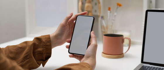 Female hands holding mock up smartphone while sitting at worktable with mock up laptop in home office