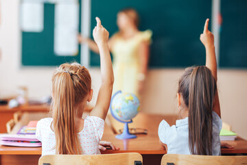 Two little schoolgirls sit at desk in school class and answer the questions of the teacher, schoolgirls raise their hands