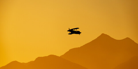 Small biplane flying at sunset silhouetted against mountains. .