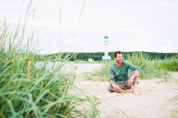 Naklejka na ściany i meble Mann , mittleren Alters sitzt am Strand , und lächelt freundlich in sich hinein. Im Hintergrund ein Leuchtturm.