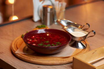 A photo of a Ukrainian borsch with a pampushka, onion, and sour cream on a wooden tray on the table in a restaurant.