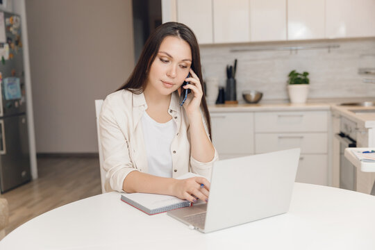 Young Woman Works In Call Center From Office Of Her Home Kitchen. Calling Customers Through Phone And Filling Out Tables On Laptop