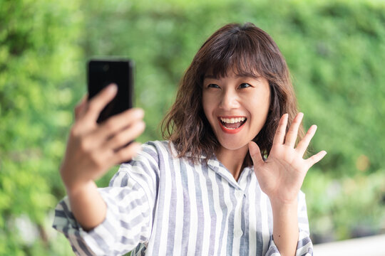 Young beautiful Asian woman using smartphone video chatting with another people. A happy girl enjoying outdoor lifestyle surrounded by greeny plant. Positive thinking concept