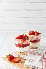 Homemade layered dessert with fresh strawberries, cream cheese or yogurt, granola and strawberry jam in glasses on white wood background. Healthy organic breakfast or snack concept. Selective focus.