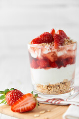 Homemade layered dessert with fresh strawberries, cream cheese or yogurt, granola and strawberry jam in glass on white wood background. Healthy organic breakfast or snack concept. Selective focus.