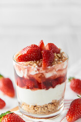 Homemade layered dessert with fresh strawberries, cream cheese or yogurt, granola and strawberry jam in glass on white wood background. Healthy organic breakfast or snack concept. Selective focus.