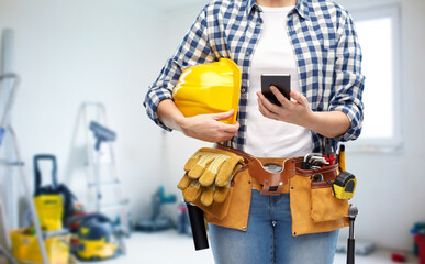 repair, construction and building concept - close up of woman or builder with smartphone, helmet and working tools on belt over utility room background