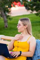 Obraz premium Young attractive girl in yellow dress works at laptop while sitting on bench in park. Concept of remote work from anywhere in the pestilence. Convenience. Freelance Worker.
