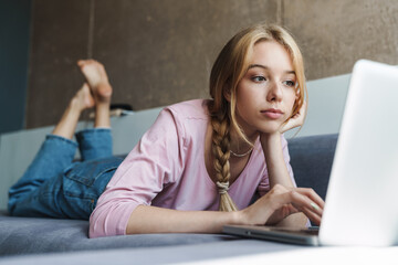 Photo of focused beautiful woman using laptop while lying on couch