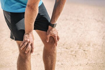 Male runner resting taking a break tired with knee pain after beach jog wearing smartwatch wearable technology for exercise monitoring weight loss.