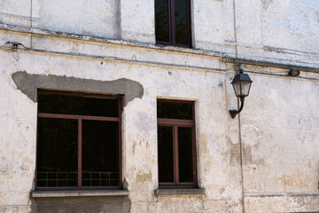 Fototapeta na wymiar Decayed exterior facade white wall with peeling plaster some wooden windows and a lantern. vintage theme background