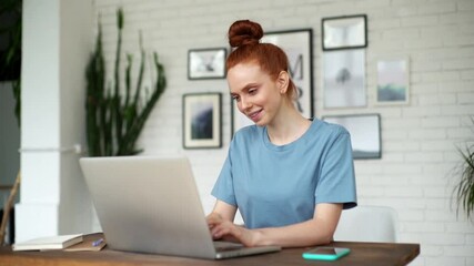 Smiling young redhead woman wearing fashion casual clothing is typing on laptop at the desk in light cozy room at the home office. Concept of remote education from home. Shooting in slow motion.