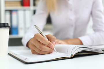 Cropped photo of lady sitting at the table at workplace while making notes in notebook