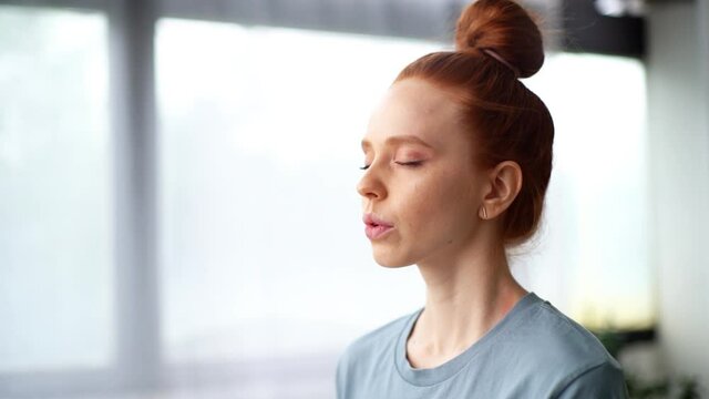 Close-up face of focused redhead young woman doing breathing yogic practices at home office. Girl with closed eyes meditating on background of large window. Cute lady is making deep breath-exhalation.