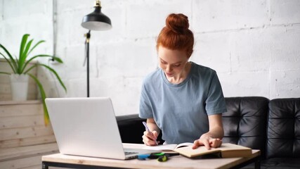Beautiful redhead young woman student is noting into the workbook important information from the book in preparation for the lessons at table with laptop. Girl doing paperwork with book in home office