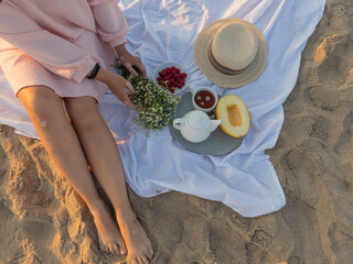 Obraz na płótnie Canvas A girl on the beach meets the dawn, female hands with vitiligo hold a bouquet of daisies, against the background of a tray with melon, raspberries and tea with daisies, romantic atmosphere
